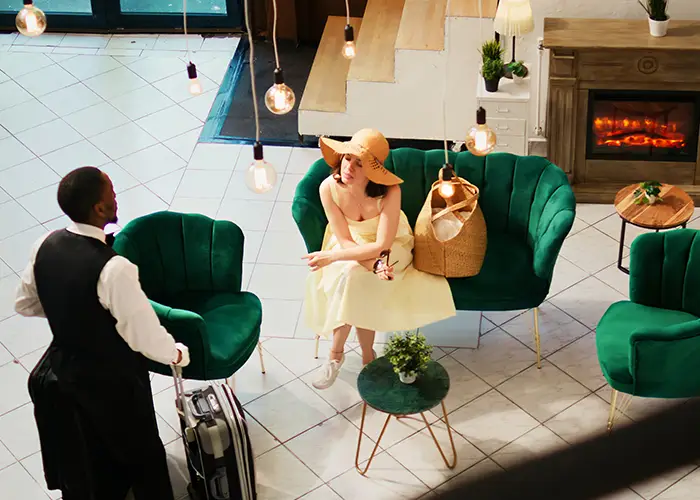Elegant lounge area with green armchairs, a fireplace, and a person waiting, with a waiter approaching in a hotel.