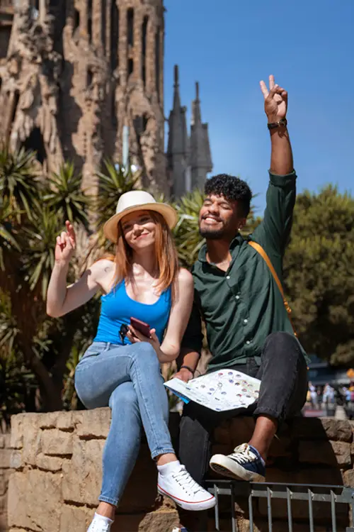 traveler couple sitting in front of Sagrada Familia, one holding a map, both making peace signs