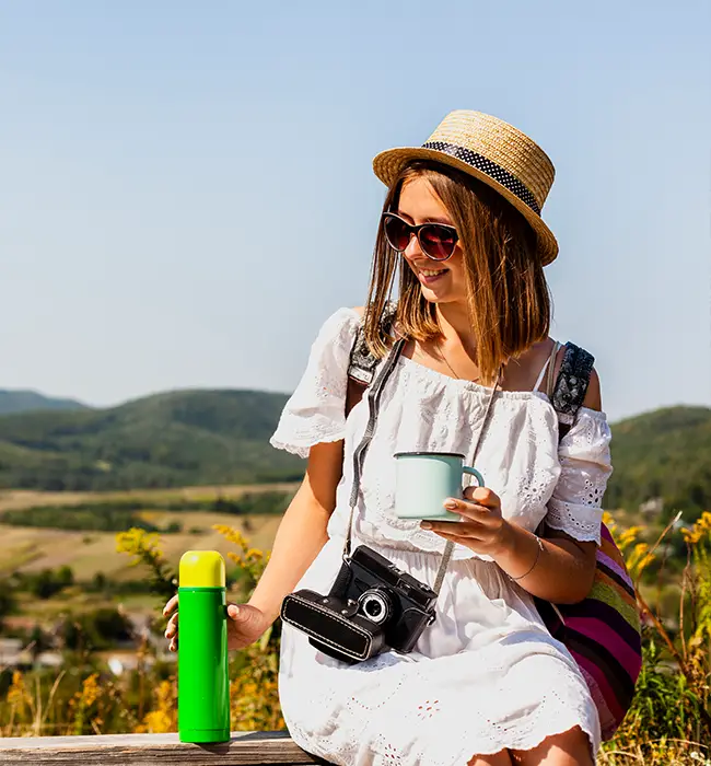 a lady traveler in a white dress with a hat holding a mug and camera, with a green flask beside, outdoor setting