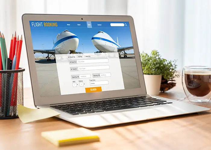 Laptop on desk displaying a flight booking website with an airplane in the background, beside a coffee cup.