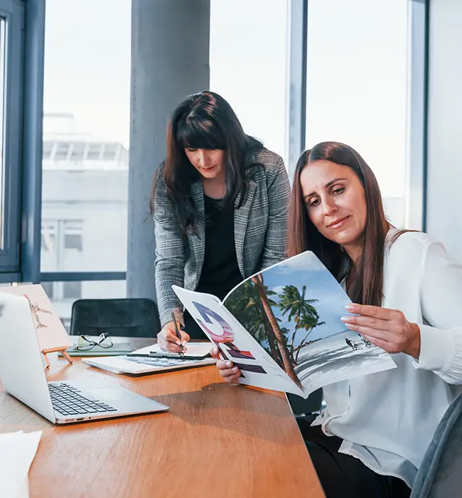 Two individuals at a table with a laptop, documents, and one holding a magazine