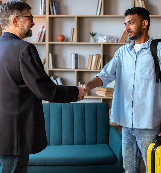 a man shaking hands with a travel agent in a room with a bookshelf and blue couch.