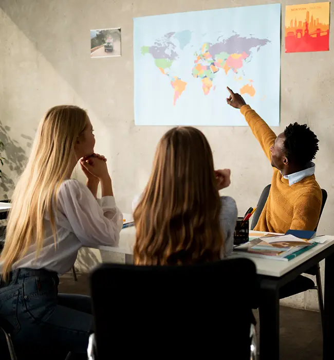 a travel agent explaining to traveler in a meeting with pointing at a world map on the wall.