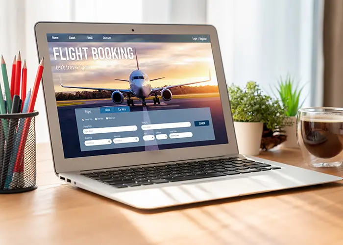 Laptop on desk with flight booking website displayed, beside a coffee cup and plant.