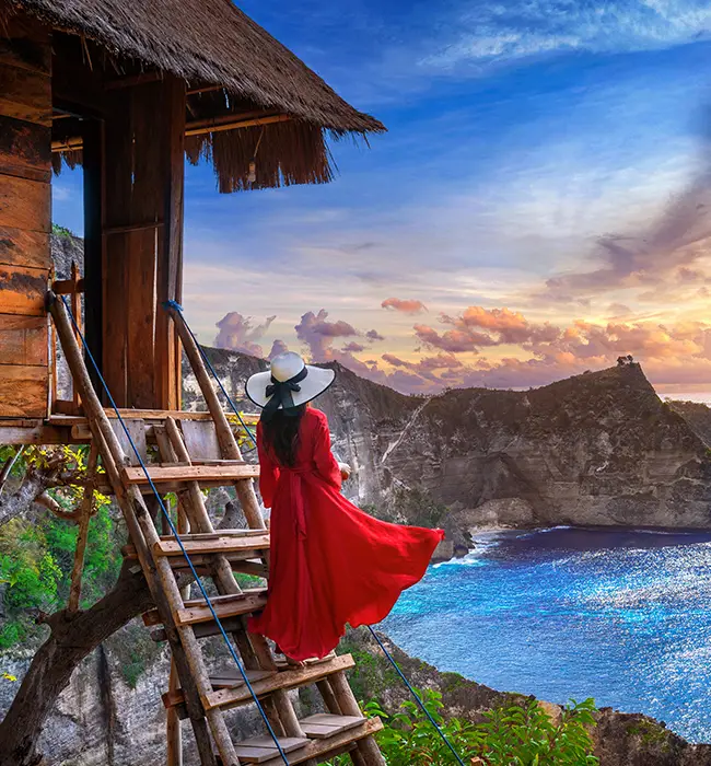 a traveler Woman in red dress on wooden stairs by a hut overlooking a coastal cliff at sunset.