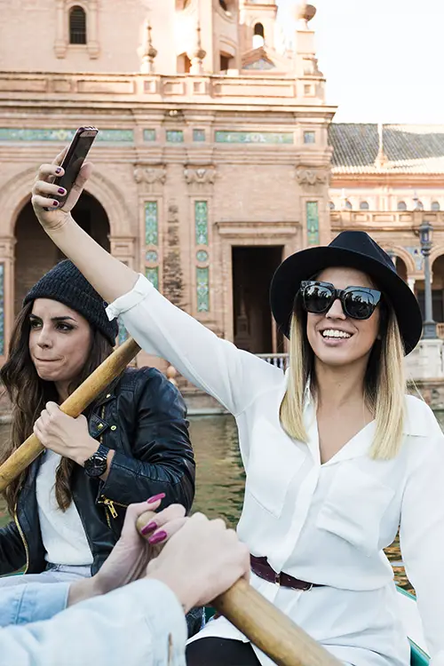 Two lady travelers in a boat, one taking a selfie, with historic architecture in the background.