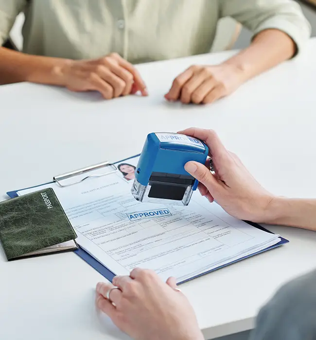 A person stamping 'APPROVED' on a visa document with another person observing.